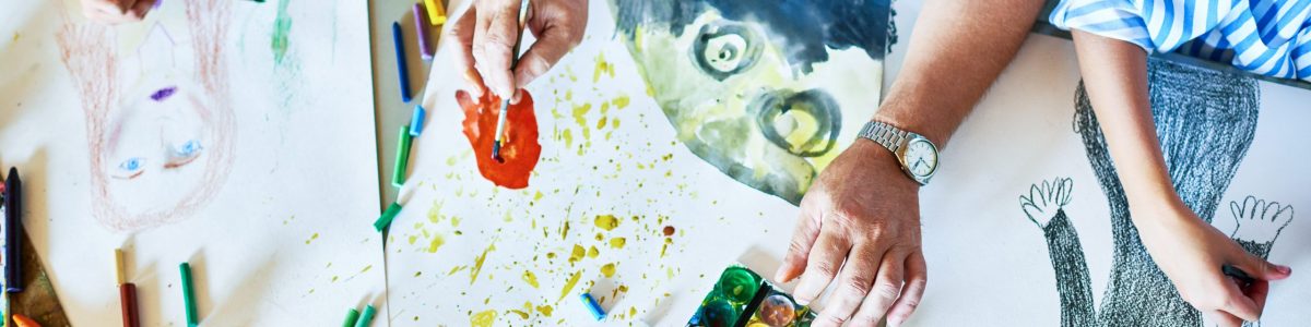 Top view of childrens drawings on table in art class with group of people painting and teachers hands